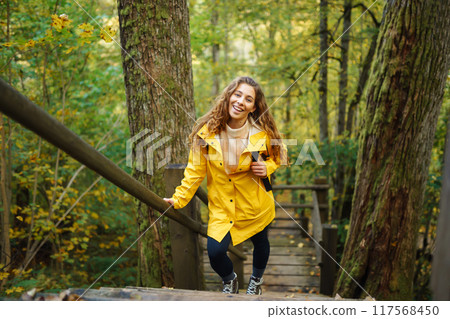Woman in a yellow coat with backpack walks along wooden staircase, enjoying autumn scenery in forest 117568450