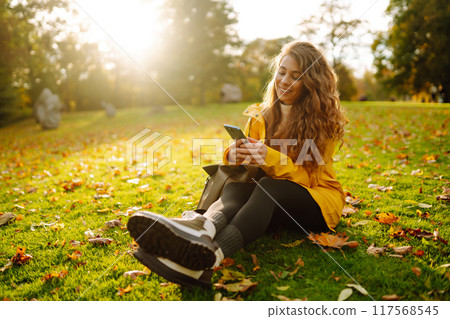 A smiling woman sits outdoors on the lawn among yellow autumn leaves with a phone in her hands. 117568545