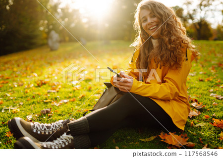 A smiling woman sits outdoors on the lawn among yellow autumn leaves with a phone in her hands. 117568546