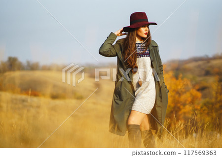 Beautiful stylish girl walks in the autumn park. The girl is dressed in a green coat and a red hat. 117569363