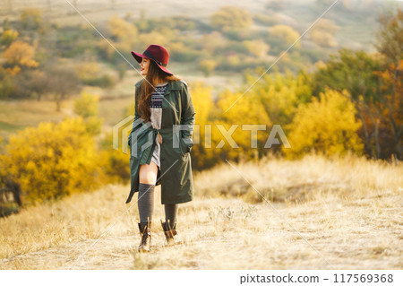 Beautiful stylish girl walks in the autumn park. The girl is dressed in a green coat and a red hat. 117569368