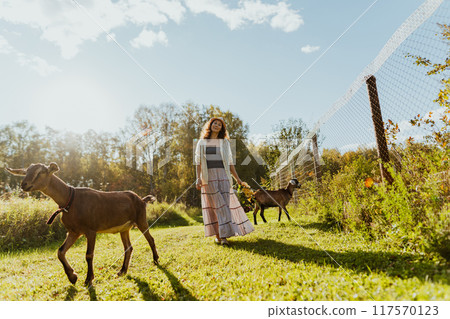 Two Nubian goats graze on a free-range pasture while a woman walks beside them. The environment is serene and eco-friendly, ideal for sustainable agriculture 117570123