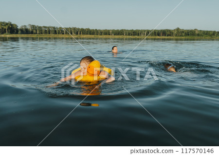 A happy children with an inflatable yellow duck is enjoying a safe swim at the lake. Their mother is watching over her, ensuring her safety and protection A happy children with an inflatable yellow duck is enjoying a safe swim at the lake. Their mother is watching over her, ensuring her safety and protection 117570146