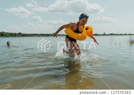 A happy child with an inflatable yellow duck is splashing in the lake on a bright summer day. She is having fun and enjoying her vacation 117570163