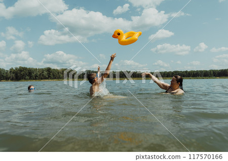 Mother and her children are enjoying a summer day at the lake, throwing an inflatable duck float. The bright sun and clear water make for a perfect vacation setting Mother and her children are enjoying a summer day at the lake, throwing an inflatable duck float. The bright sun and clear water make for a perfect vacation setting 117570166