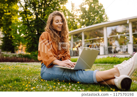 Portrait of young beautiful female freelancer sitting on green meadow with laptop. Education online. Portrait of young beautiful female freelancer sitting on green meadow with laptop. Education online. 117570198
