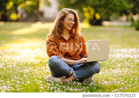 Portrait of young beautiful female freelancer sitting on green meadow with laptop. Education online. Portrait of young beautiful female freelancer sitting on green meadow with laptop. Education online. 117570225