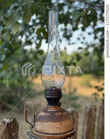 A kerosene lamp is placed on a dilapidated wooden fence. Against a backdrop of the forest. The background is blurred A kerosene lamp is placed on a dilapidated wooden fence. Against a backdrop of the forest. The background is blurred 117570428