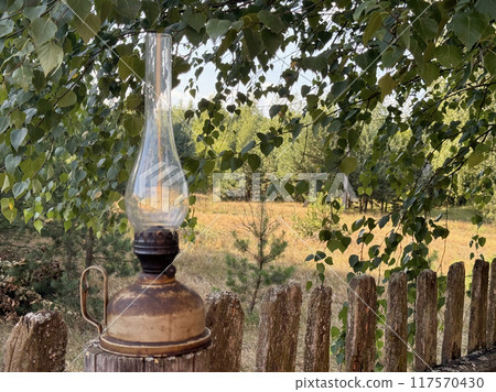 A kerosene lamp is placed on a dilapidated wooden fence. Against a backdrop of the forest. A kerosene lamp is placed on a dilapidated wooden fence. Against a backdrop of the forest. 117570430