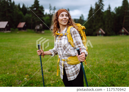 Stylish woman traveler with yellow hiking backpack and hiking stiks enjoys scenery. Active lifestyle 117570942