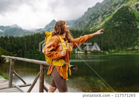 Female traveler with yellow hiking backpack against the backdrop of mountain lake with hiking poles. 117571265