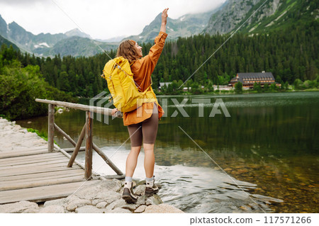 Female traveler with yellow hiking backpack against the backdrop of mountain lake with hiking poles. Female traveler with yellow hiking backpack against the backdrop of mountain lake with hiking poles. 117571266