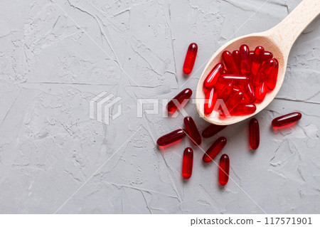 Vitamin capsules in a spoon on a colored background. Pills served as a healthy meal. Red soft gel vitamin supplement capsules on spoon 117571901