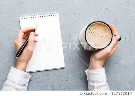 Woman hand with pencil writing on notebook and hold coffee cup. Woman working on office table with coffee Woman hand with pencil writing on notebook and hold coffee cup. Woman working on office table with coffee 117572014