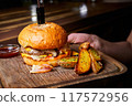 Burger with beef cutlet, a wheat bun and cheese, with a knife stuck into it, served with baked potatoes 117572956