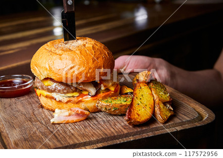 Burger with beef cutlet, a wheat bun and cheese, with a knife stuck into it, served with baked potatoes Burger with beef cutlet, a wheat bun and cheese, with a knife stuck into it, served with baked potatoes 117572956