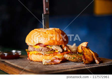 Burger with beef cutlet, a wheat bun and cheese, with a knife stuck into it, served with baked potatoes Burger with beef cutlet, a wheat bun and cheese, with a knife stuck into it, served with baked potatoes 117572961
