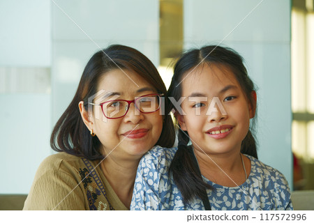 Portrait Asian mother and daughter sitting together, healthy middle- aged woman and child at 9 to 10 years old, both looking at camera, indoors image with natural light. 117572996