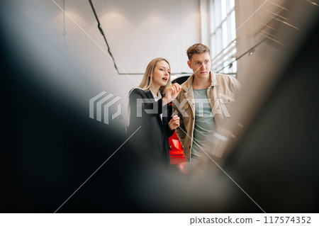 View through door vehicle to professional female car dealer in business suit telling male buyer about luxurious car in dealership office. Doubtful man client looking at auto in motor show. 117574352