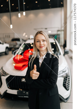 Vertical portrait of successful salesperson female in business suit showing thumb up gesture, smiling looking at camera standing at modern salon on background of new white car. Concept choosing auto 117574370