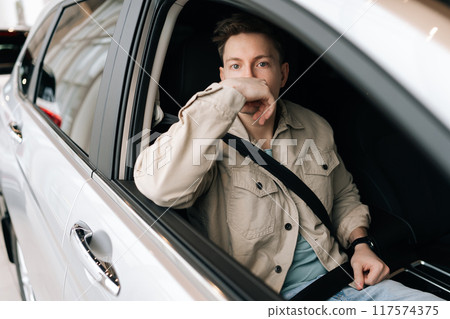 Portrait of pensive passenger male sitting on front seat fastening belt harness, looking at camera. Front view of young man with buckled security belt in car. Concept of safety lifestyle. 117574375