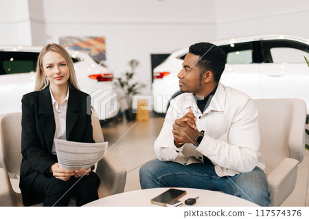 Portrait of female car dealer consulting serious black buyer, giving purchase agreement for review at showroom. African American customer receiving car purchase contract before signing in dealership 117574376