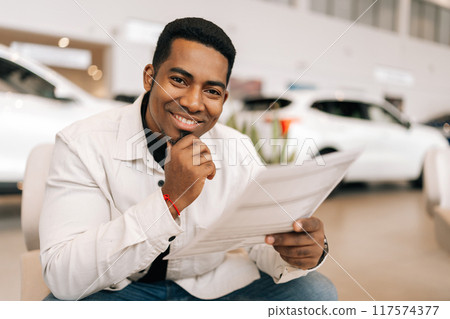 Portrait of cheerful African male buyer reading car purchase agreement before signing in dealership office, looking at camera with happy expression. Concept of choosing and buying new auto at showroom 117574377