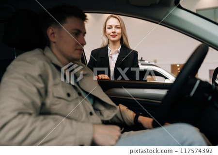 View through side window of car to professional sales female manager in suit talking to male client sitting in car in showroom choosing automobile holding steering wheel. Concept of buying new auto. View through side window of car to professional sales female manager in suit talking to male client sitting in car in showroom choosing automobile holding steering wheel. Concept of buying new auto. 117574392
