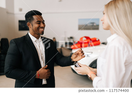 Side view of cheerful smiling female buyer getting car key and congratulation from black auto dealer male while buying new auto in dealership. Woman car owner during handing over keys at showroom. 117574400