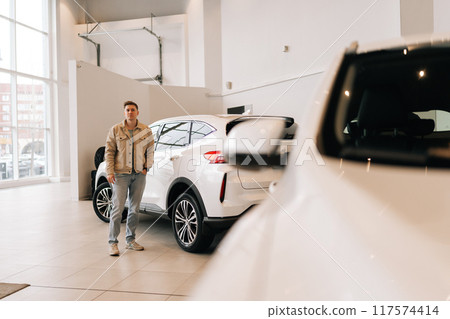 Remote view of pensive young man choosing new automobile walking around luxury cars in car dealership. Doubtful customer male walking in salesroom and studies transportation models. 117574414