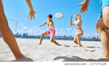 Dynamic image of young people, men and women, friends playing frisbee game in beach by the river under clear blue sky 117575220