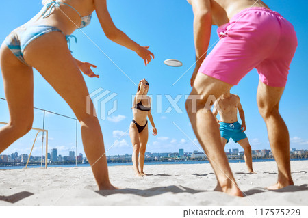Dynamic image of young people, men and women, friends playing frisbee game in beach by the river under clear blue sky 117575229