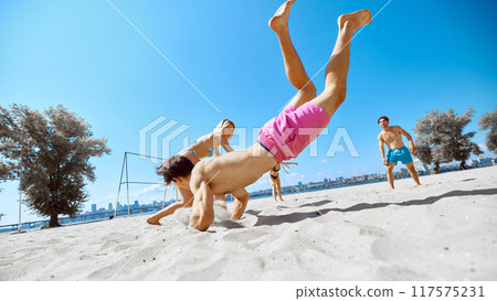 Dynamic game. Positive and active young people, friends gathering on beach by the river and playing energetic frisbee game 117575231