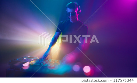 DJ in headphones immersed in of vibrant lights, mixing beats as colors pulse around him, creating an electrifying atmosphere. DJ in headphones immersed in of vibrant lights, mixing beats as colors pulse around him, creating an electrifying atmosphere. 117575344