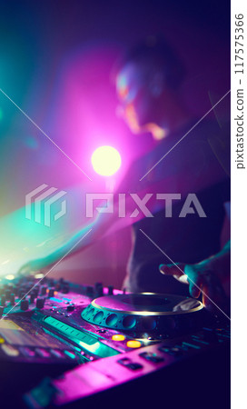 DJ, immersed in vibrant hues, crafts musical journey, his hands moving effortlessly over controls as lights dance around him. DJ, immersed in vibrant hues, crafts musical journey, his hands moving effortlessly over controls as lights dance around him. 117575366
