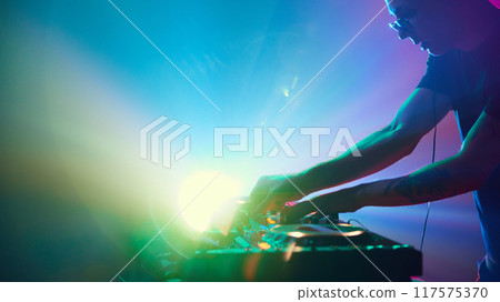 DJ stands in vibrant spotlight, surrounded by vibrant light show, his music shaping electric energy of scene. 117575370