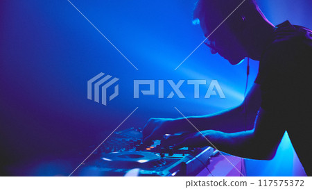 Symphony of light and sound. DJ commands deck, his movements synchronized with shifting spotlights that paint dancefloor. 117575372