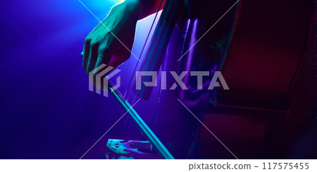 Hand elegantly holding bow and slightly slides over double bass strings in dramatic stage smoke and vibrant neon light. Hand elegantly holding bow and slightly slides over double bass strings in dramatic stage smoke and vibrant neon light. 117575455