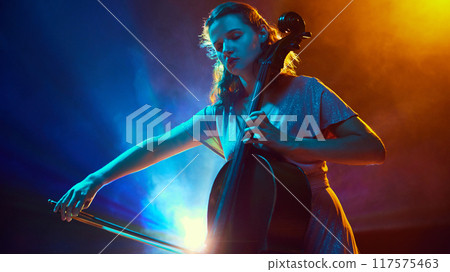 Young talented woman, musician, performing solo cello on stage surrounded dramatic smoke and neon light. Orchestra. Young talented woman, musician, performing solo cello on stage surrounded dramatic smoke and neon light. Orchestra. 117575463