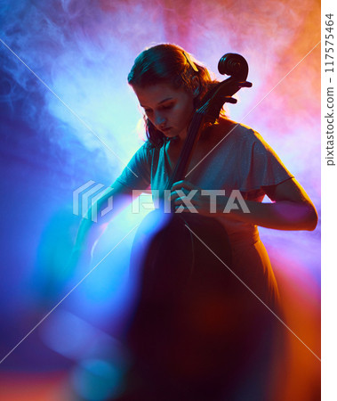 Young talented woman, musician, playing cello on stage surrounded dramatic smoke and neon light. Orchestra. Young talented woman, musician, playing cello on stage surrounded dramatic smoke and neon light. Orchestra. 117575464