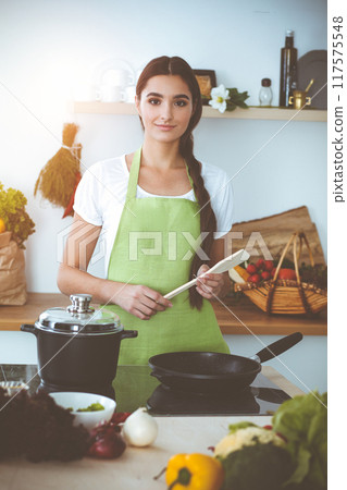 An attractive young dark-haired woman frying meal at worm pan by new keto recipe while standing and smiling in sunny kitchen. Cooking and householding concepts 117575548