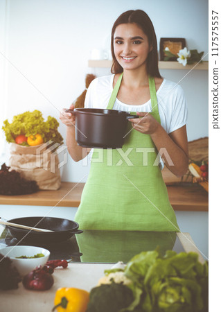 An attractive young dark-haired woman preparing soup by new keto recipe while standing and smiling in sunny kitchen. Cooking and householding concepts An attractive young dark-haired woman preparing soup by new keto recipe while standing and smiling in sunny kitchen. Cooking and householding concepts 117575557