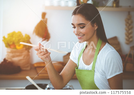 An attractive young dark-haired woman tasting ready hot meal with a wooden spoon while standing and smiling in sunny kitchen. Cooking and householding concepts An attractive young dark-haired woman tasting ready hot meal with a wooden spoon while standing and smiling in sunny kitchen. Cooking and householding concepts 117575558