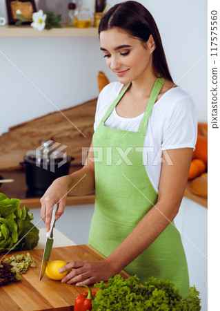 An attractive young dark-haired woman preparing delicious fresh vegetarian salad while standing and smiling near the kitchen desk. Cooking and householding concepts 117575560