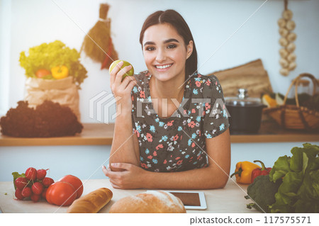 An attractive young dark-haired woman holding green apple and choosing the recipe for a delicious meal while smiling in sunny kitchen. Tablet pc is the best cookbook An attractive young dark-haired woman holding green apple and choosing the recipe for a delicious meal while smiling in sunny kitchen. Tablet pc is the best cookbook 117575571