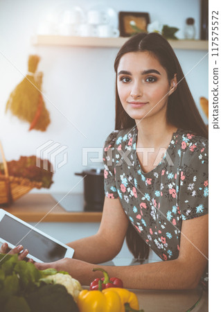 An attractive young dark-haired woman chooses the recipe for a delicious meal while sitting at the table in sunny kitchen An attractive young dark-haired woman chooses the recipe for a delicious meal while sitting at the table in sunny kitchen 117575572