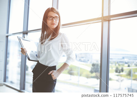 Beautiful female specialist with clipboard standing in sunny office and smiling charmingly. Business people concept Beautiful female specialist with clipboard standing in sunny office and smiling charmingly. Business people concept 117575745