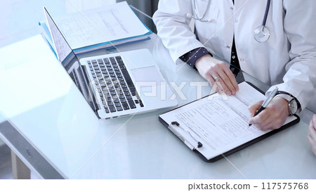 Doctor and patient are talking in clinic after usual health check up. Female physician is making notes and discussing something with a senior man. Medicine concept 117575768