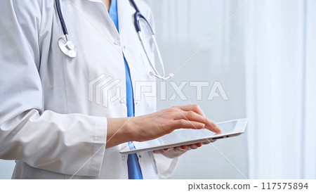 Female doctor using digital tablet in clinic. Healthcare professional with a stethoscope around her neck operating a touch screen computer while standing straight. Medicine and healthcare Female doctor using digital tablet in clinic. Healthcare professional with a stethoscope around her neck operating a touch screen computer while standing straight. Medicine and healthcare 117575894