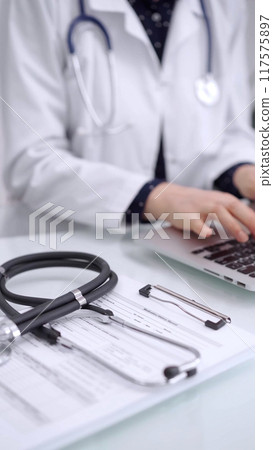 Stethoscope is lying on the glass table while doctor woman is working with laptop computer on the background, close-up. Medicine and pharmacy concept Stethoscope is lying on the glass table while doctor woman is working with laptop computer on the background, close-up. Medicine and pharmacy concept 117575897